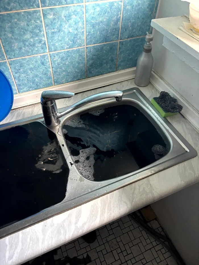Un évier de cuisine rempli d'eau sombre et de bulles, à côté d'un distributeur de savon argenté et d'une éponge verte munie d'une brosse à récurer noire, sur un plan de travail de couleur claire.