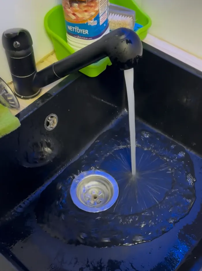 Un évier de cuisine noir d'où coule de l'eau d'un robinet noir, avec, à l'arrière-plan, un bac vert contenant une bouteille de liquide vaisselle et une brosse.