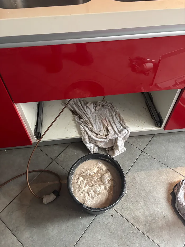 Un seau contenant un liquide mousseux posé sur un sol carrelé, sous un meuble de cuisine aux portes rouges, avec à l'intérieur du meuble un chiffon sale et un tuyau débranché.